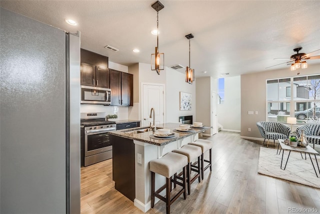 kitchen with a center island with sink, sink, dark brown cabinets, dark stone counters, and appliances with stainless steel finishes