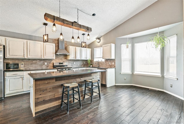 kitchen with stainless steel appliances, vaulted ceiling, dark countertops, wall chimney range hood, and a center island