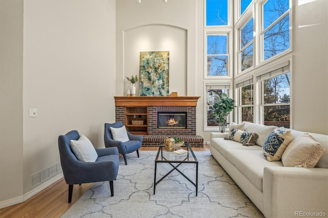 living room featuring wood finished floors, visible vents, baseboards, a towering ceiling, and a brick fireplace