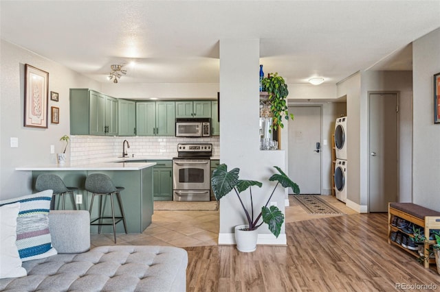 kitchen with stacked washer and dryer, appliances with stainless steel finishes, backsplash, green cabinetry, and kitchen peninsula