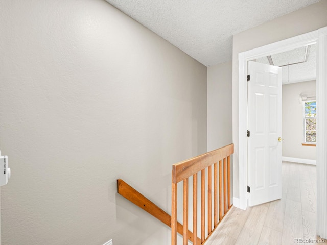 corridor featuring an upstairs landing, baseboards, light wood finished floors, a textured ceiling, and attic access