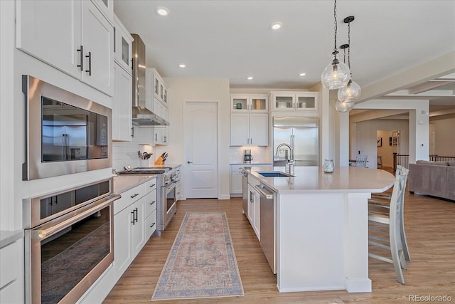 kitchen with a sink, light wood-style floors, wall chimney exhaust hood, light countertops, and built in appliances