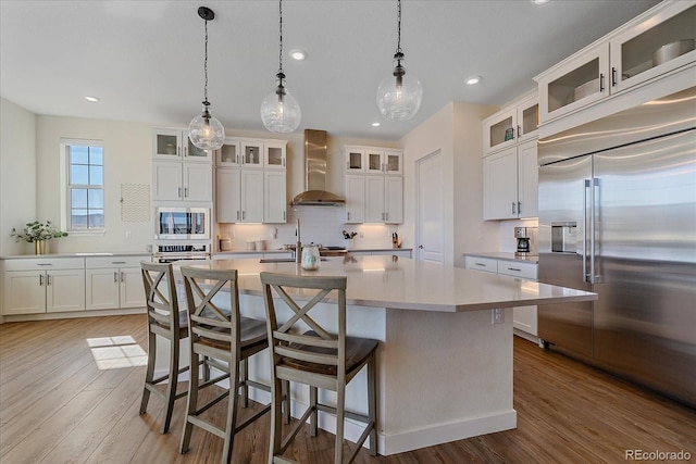 kitchen with wall chimney range hood, built in appliances, light countertops, decorative backsplash, and wood finished floors