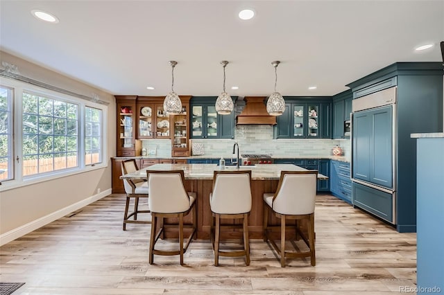 kitchen with blue cabinetry, hanging light fixtures, light stone counters, an island with sink, and decorative backsplash