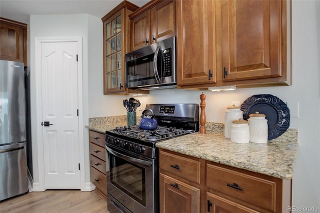 kitchen featuring appliances with stainless steel finishes, light wood-style floors, glass insert cabinets, and light stone countertops