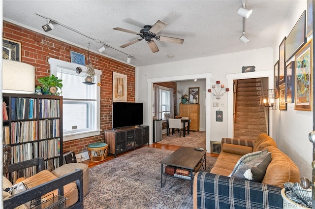 living room featuring a textured ceiling, hardwood / wood-style flooring, rail lighting, ceiling fan, and brick wall