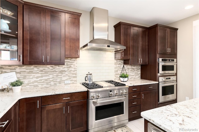 kitchen with light tile patterned flooring, stainless steel appliances, wall chimney range hood, backsplash, and light stone countertops