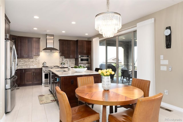 kitchen with stainless steel appliances, wall chimney range hood, decorative light fixtures, sink, and a kitchen island with sink