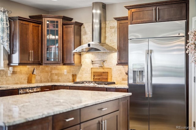 17102 W 85th Pl, Arvada, CO 80007 - kitchen featuring stainless steel appliances, wall chimney range hood, light stone countertops, glass insert cabinets, and backsplash
