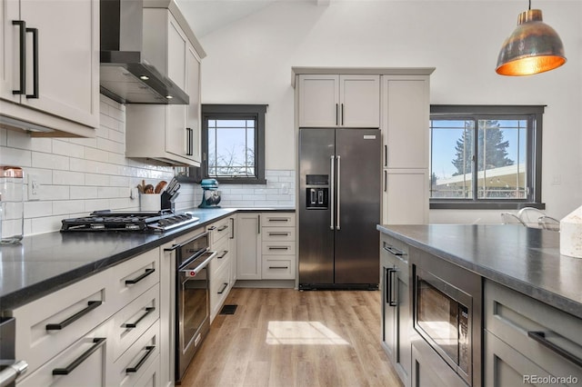 kitchen featuring backsplash, vaulted ceiling, appliances with stainless steel finishes, light wood-style floors, and wall chimney exhaust hood