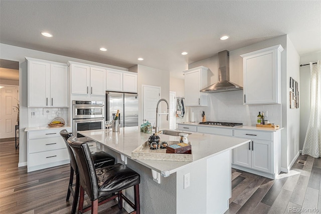 kitchen featuring stainless steel appliances, dark wood-style flooring, wall chimney exhaust hood, and a center island with sink
