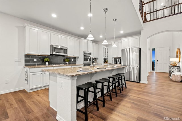 kitchen featuring arched walkways, stainless steel appliances, light wood-style floors, a kitchen island with sink, and white cabinetry