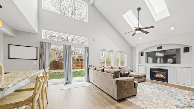 living room featuring high vaulted ceiling, a fireplace, a skylight, light wood-type flooring, and ceiling fan
