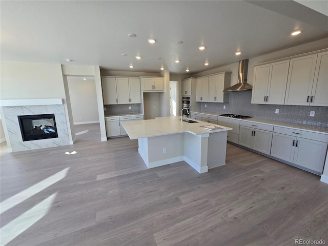 kitchen featuring wall chimney range hood, gas cooktop, light hardwood / wood-style flooring, an island with sink, and a fireplace