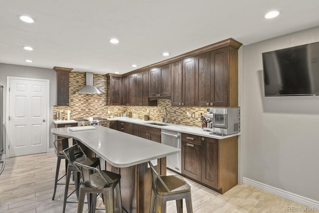 kitchen featuring sink, wall chimney exhaust hood, appliances with stainless steel finishes, dark brown cabinets, and a breakfast bar area