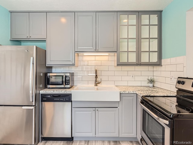 kitchen with gray cabinets, decorative backsplash, appliances with stainless steel finishes, glass insert cabinets, and a sink