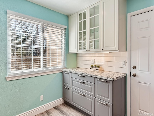 kitchen with gray cabinetry, baseboards, light wood-style floors, backsplash, and glass insert cabinets