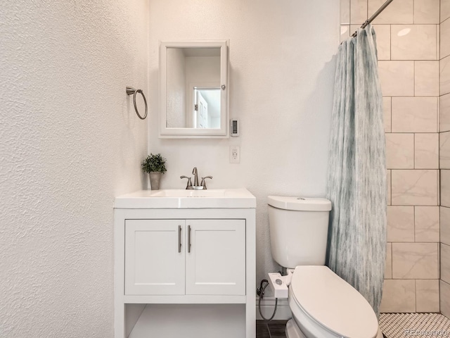 bathroom featuring toilet, a textured wall, tiled shower, and vanity