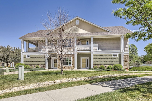 view of front of home featuring stone siding, a balcony, roof with shingles, and a front lawn