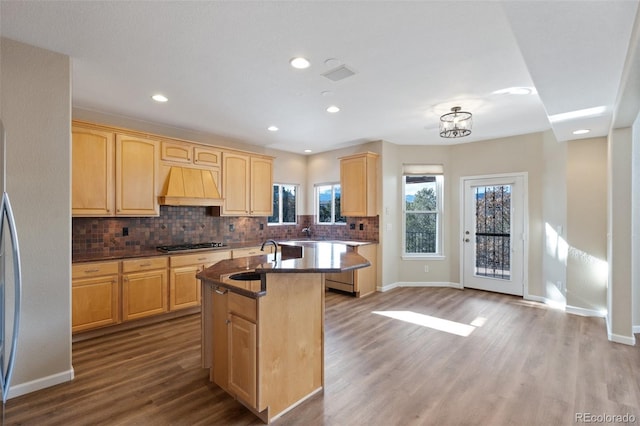 kitchen featuring sink, custom exhaust hood, stainless steel gas stovetop, light brown cabinetry, and a center island with sink