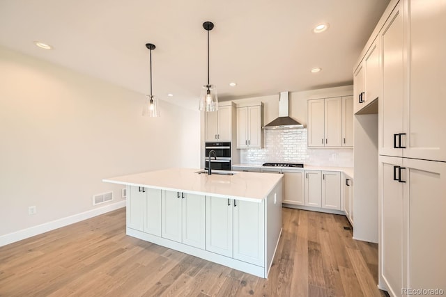 kitchen featuring pendant lighting, wall chimney range hood, an island with sink, and white cabinets