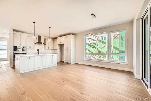 kitchen featuring white cabinetry, a center island with sink, pendant lighting, decorative backsplash, and wall chimney range hood