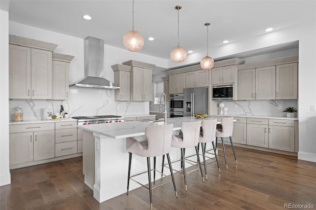 kitchen with appliances with stainless steel finishes, a breakfast bar, hanging light fixtures, a center island with sink, and wall chimney exhaust hood