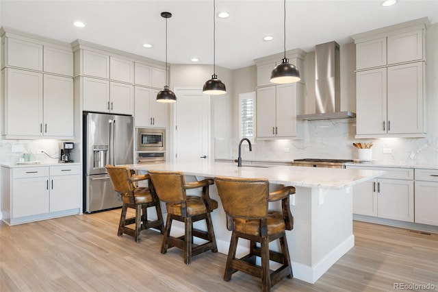 kitchen with an island with sink, stainless steel appliances, a breakfast bar, wall chimney exhaust hood, and hanging light fixtures