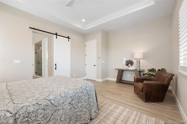 bedroom featuring a barn door, light wood-type flooring, ceiling fan, a tray ceiling, and crown molding