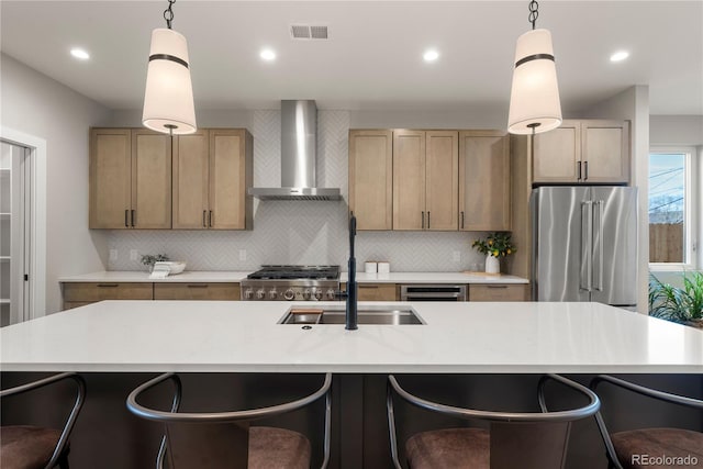 kitchen with appliances with stainless steel finishes, light brown cabinets, decorative light fixtures, and wall chimney range hood