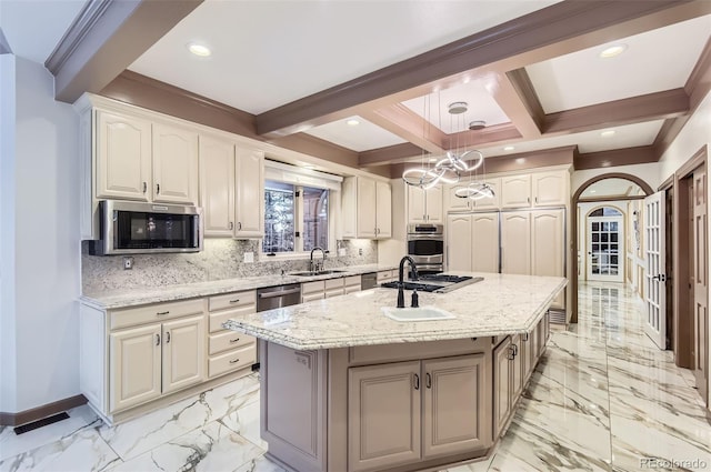 kitchen with appliances with stainless steel finishes, an island with sink, pendant lighting, light stone countertops, and decorative backsplash