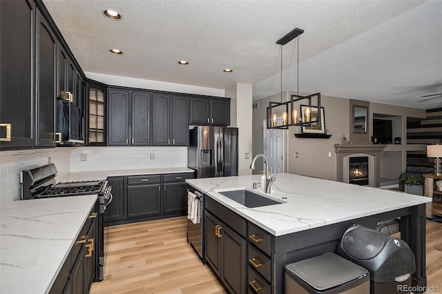 kitchen with a kitchen island with sink, light wood-style flooring, a sink, tasteful backsplash, and appliances with stainless steel finishes