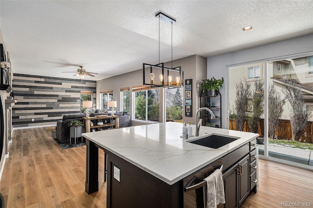 kitchen with light wood finished floors, a sink, a textured ceiling, a center island with sink, and light stone countertops