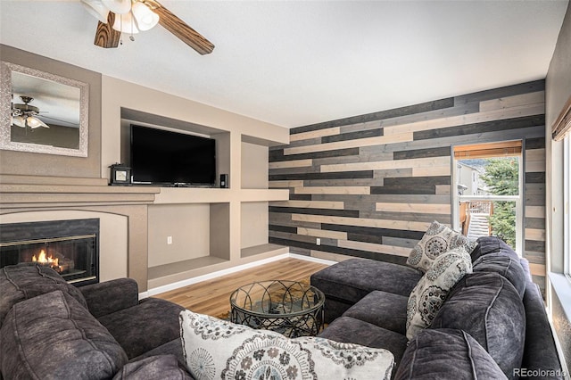 living room featuring wood finished floors, a glass covered fireplace, a ceiling fan, and wooden walls