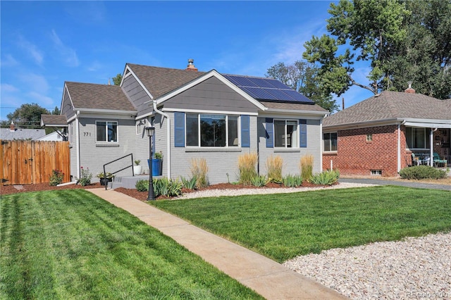 view of front facade with brick siding, a front yard, roof mounted solar panels, roof with shingles, and fence