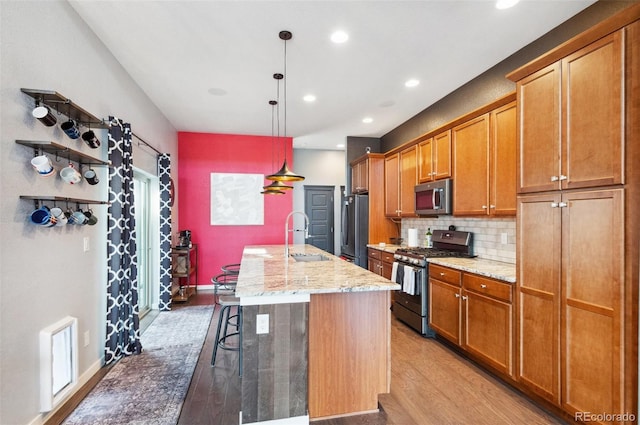 kitchen with appliances with stainless steel finishes, decorative light fixtures, light stone counters, a center island with sink, and light hardwood / wood-style floors