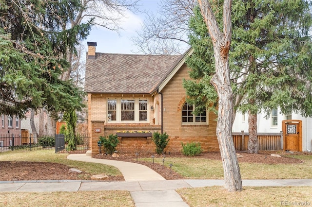 tudor house featuring brick siding, a shingled roof, a chimney, and fence