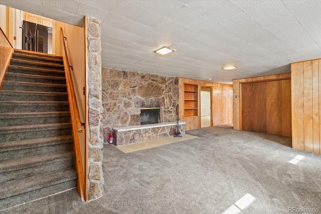 unfurnished living room with a stone fireplace, stairway, wooden walls, carpet floors, and built in shelves