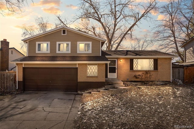 tri-level home featuring driveway, fence, roof with shingles, an attached garage, and brick siding