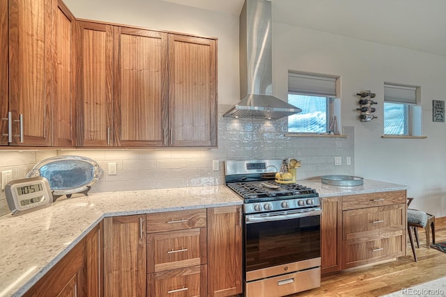 kitchen with wall chimney range hood, stainless steel range with gas stovetop, light hardwood / wood-style floors, light stone countertops, and decorative backsplash