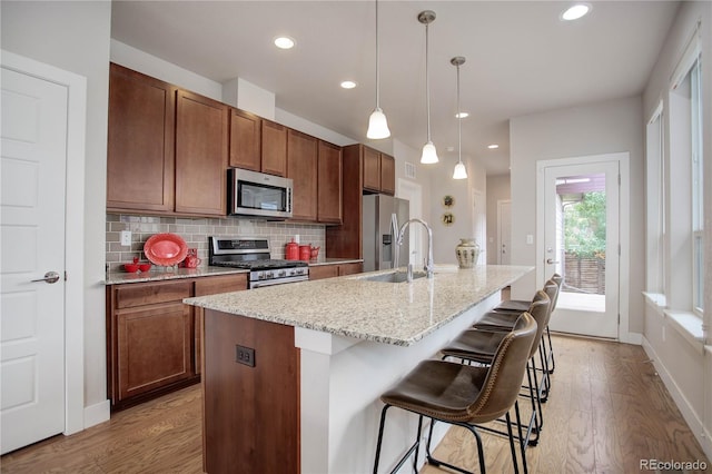 kitchen featuring light hardwood / wood-style floors, appliances with stainless steel finishes, tasteful backsplash, decorative light fixtures, and an island with sink