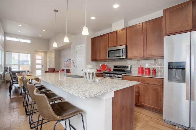 kitchen with a kitchen island with sink, backsplash, pendant lighting, light wood-type flooring, and appliances with stainless steel finishes