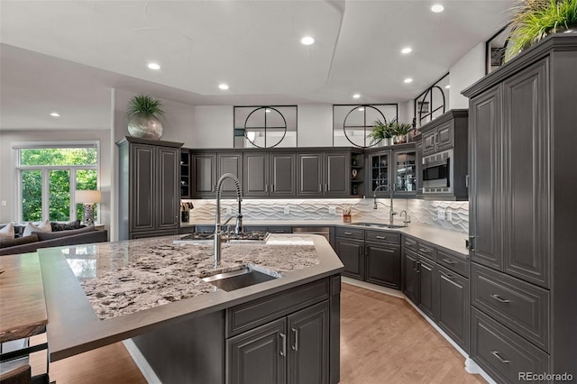 kitchen featuring sink, light stone counters, an island with sink, light hardwood / wood-style floors, and decorative backsplash