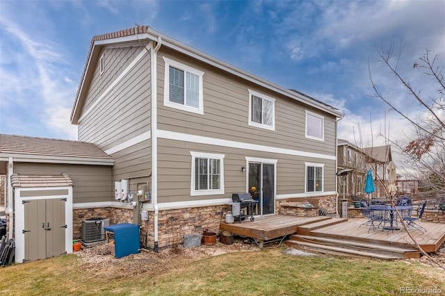 back of property featuring a deck, central air condition unit, stone siding, a lawn, and outdoor dining space