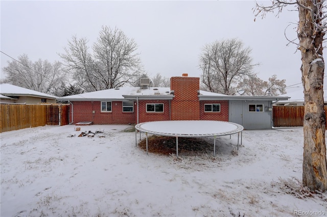 snow covered rear of property featuring brick siding, a trampoline, a chimney, and fence