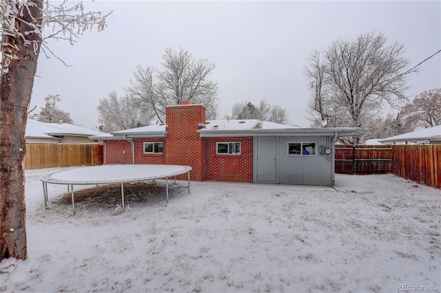 snow covered back of property with a trampoline, a fenced backyard, brick siding, and a chimney