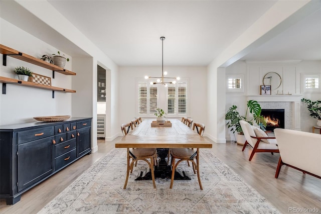 dining space featuring light wood finished floors, beverage cooler, a chandelier, a warm lit fireplace, and baseboards