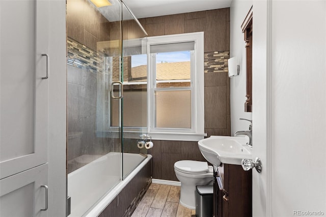 full bathroom featuring toilet, hardwood / wood-style floors, combined bath / shower with glass door, vanity, and wood walls