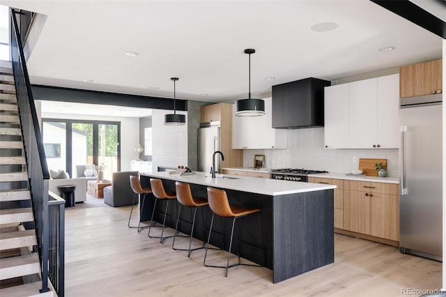 kitchen with white cabinets, a center island with sink, wall chimney exhaust hood, appliances with stainless steel finishes, and decorative light fixtures