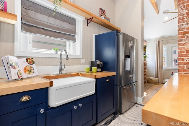 kitchen featuring wooden counters, blue cabinets, a sink, stainless steel fridge, and brick wall